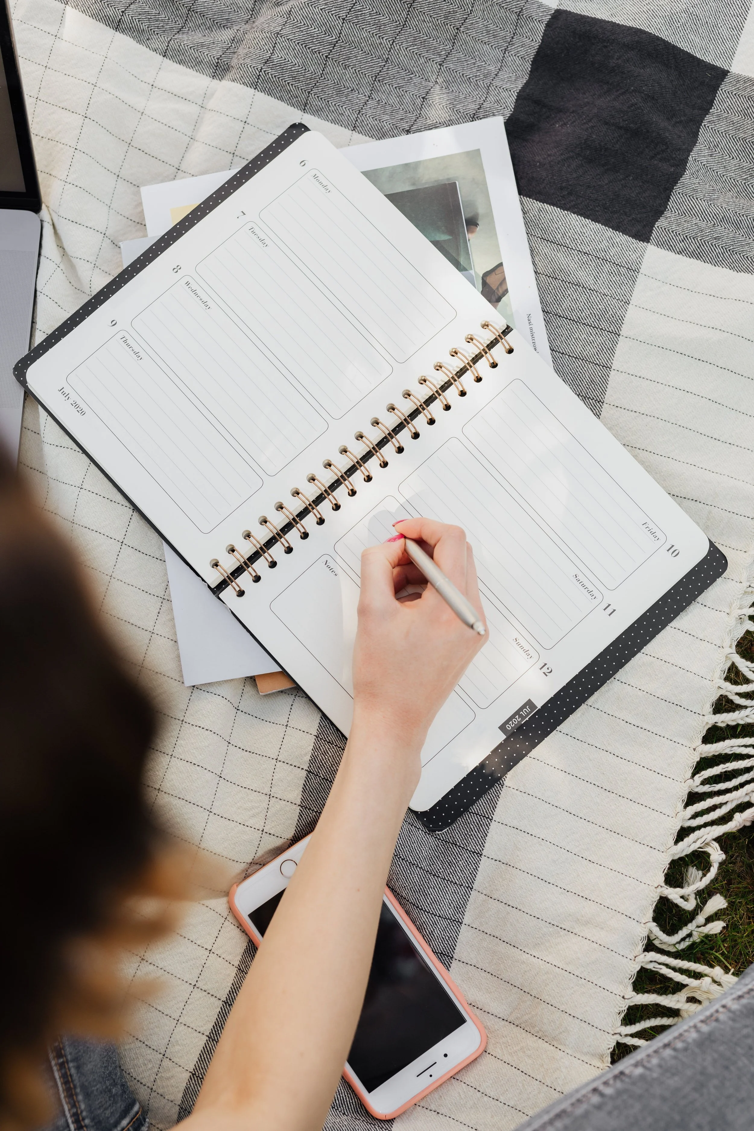 A person writing in a weekly planner with a silver pen. The planner is open on a checkered fabric surface, and there is a smartphone with a pink case and a laptop partially visible nearby.