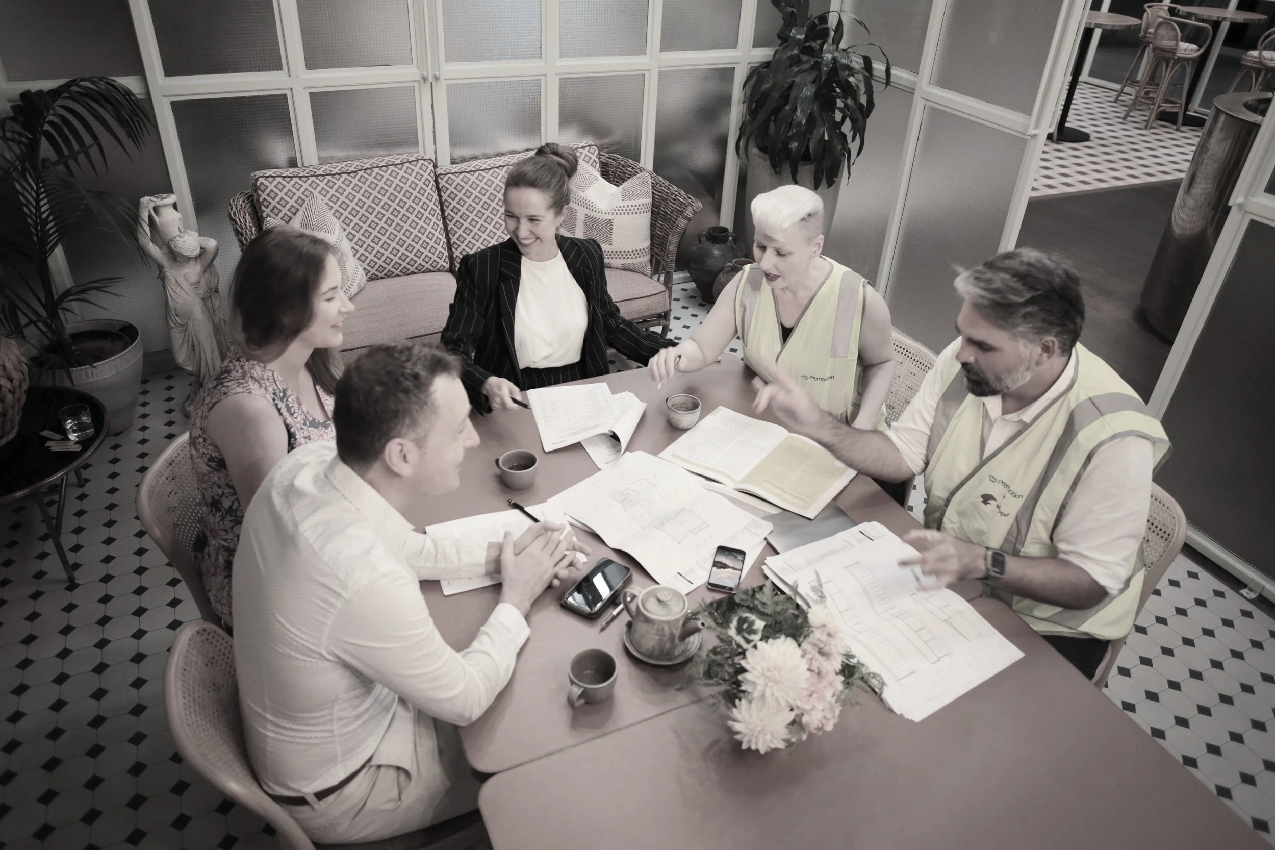 Group of five people sitting around a table, having a meeting, with plans and documents, in a well-lit room with patterned tiles and decorative plants.