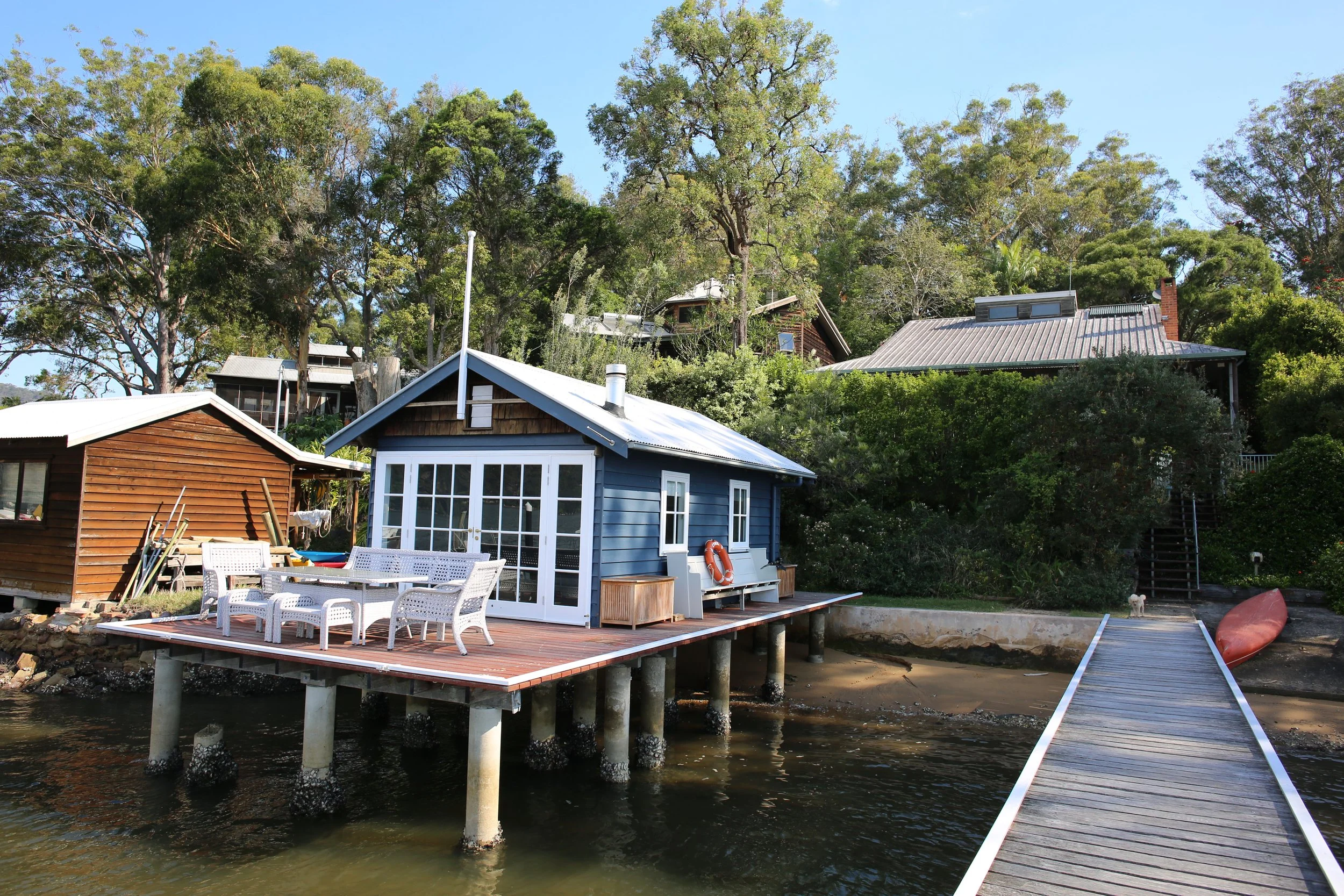 Waterfront house with a blue exterior and a deck with outdoor furniture, connected by a wooden walkway to a sandy shore, surrounded by trees and other houses.