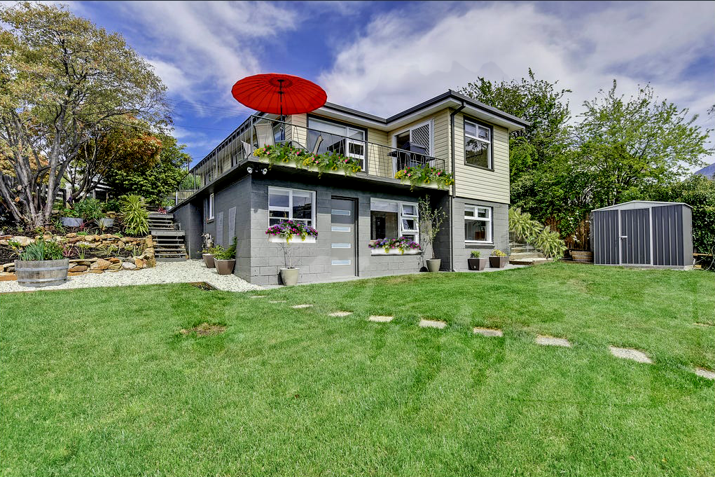 A two-story house with a gray base and beige upper level, surrounded by a green lawn and trees, featuring a red patio umbrella on a balcony and flower pots on windowsills.