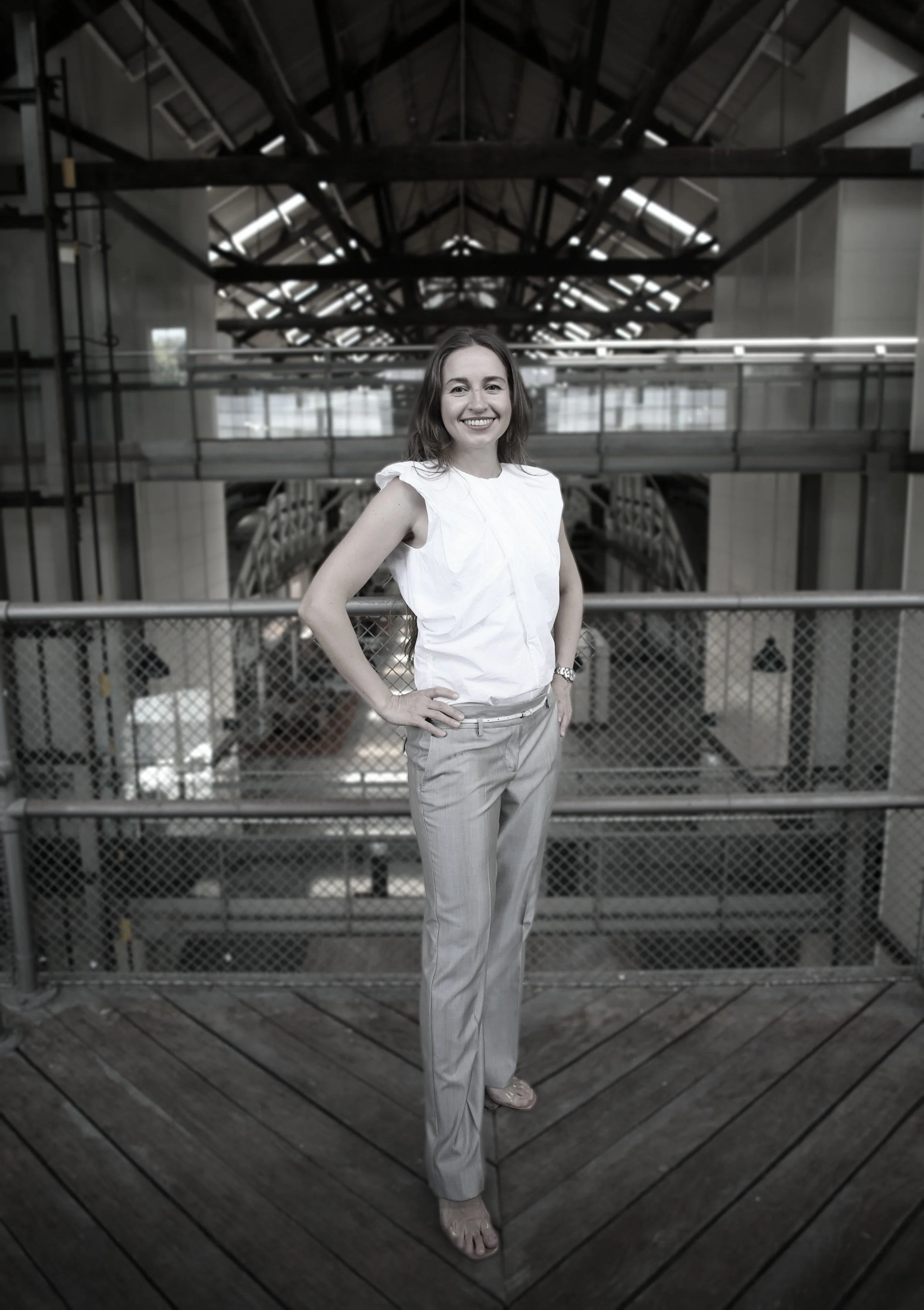 A woman standing on a wooden platform in an industrial-style building, smiling at the camera. She is wearing a white sleeveless top and light-colored striped pants.