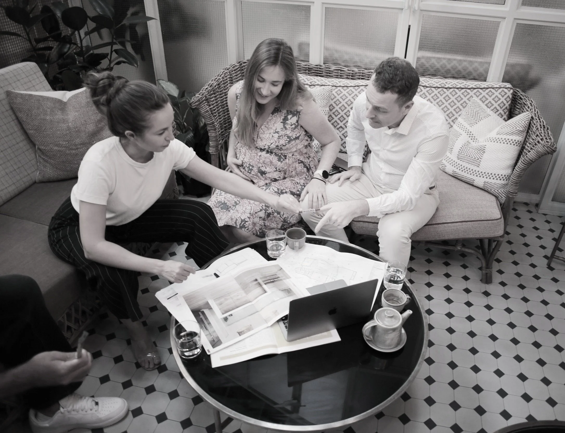 Four people sitting around a table, engaged in a discussion or collaborative work. There are papers, a laptop, and glasses of water on the table, with a cozy indoor setting and a patterned tiled floor.