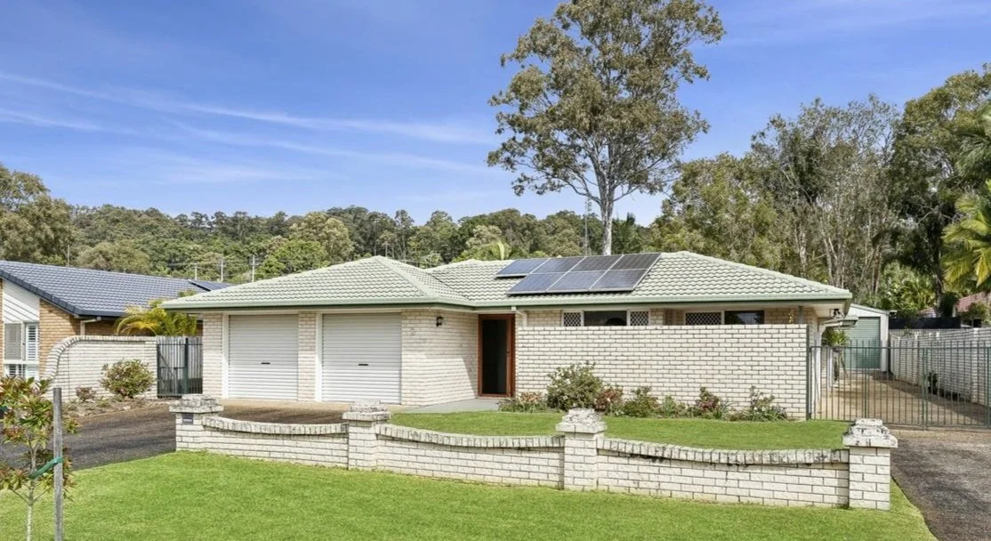 Single-story house with white brick exterior, green tiled roof with solar panels, front yard with lawn and small garden, driveway and garage, set in a suburban area with trees and greenery in the background.