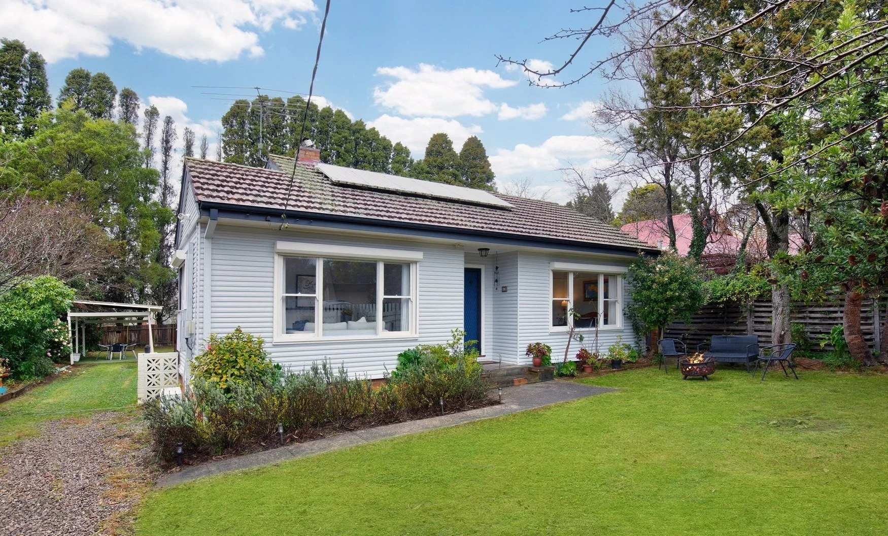 A white house with a blue door and solar panels on the roof, surrounded by a green lawn, trees, and outdoor furniture, under a partly cloudy sky.