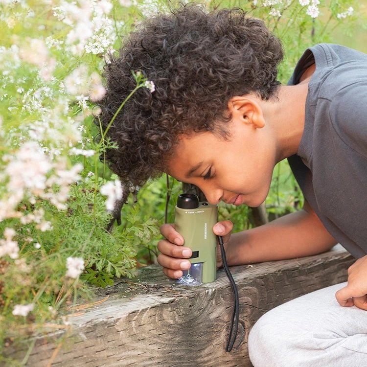 Image of a boy using a pocket microscope outside on a wooden beam in a grassy area