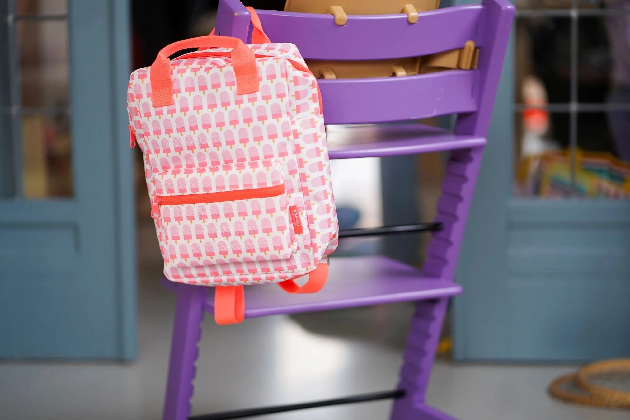Image of a neon pink backpack with an ice cream pop hanging on a purple wooden chair.