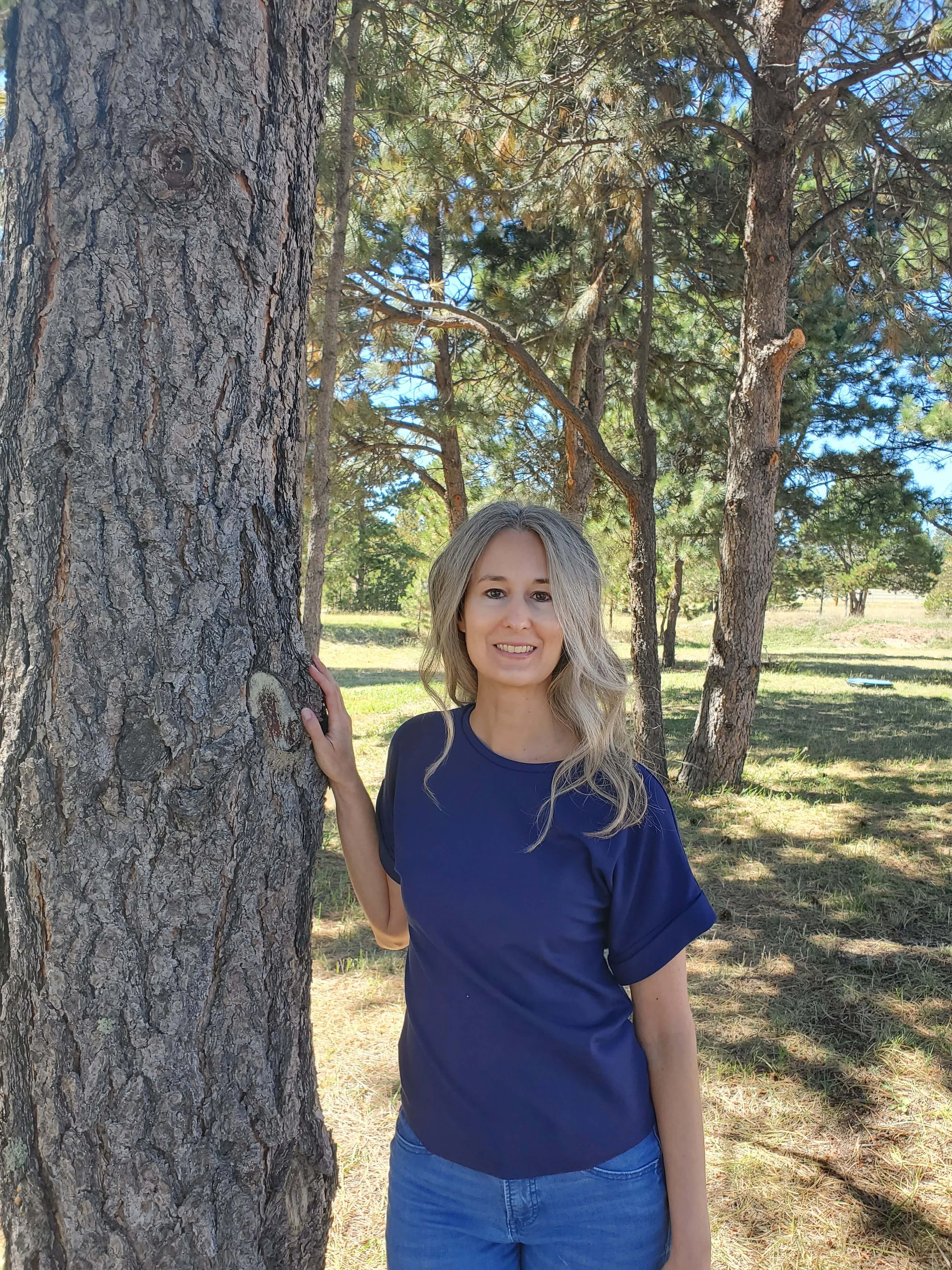 Person standing next to a tree in a wooded area, wearing a blue shirt and jeans.