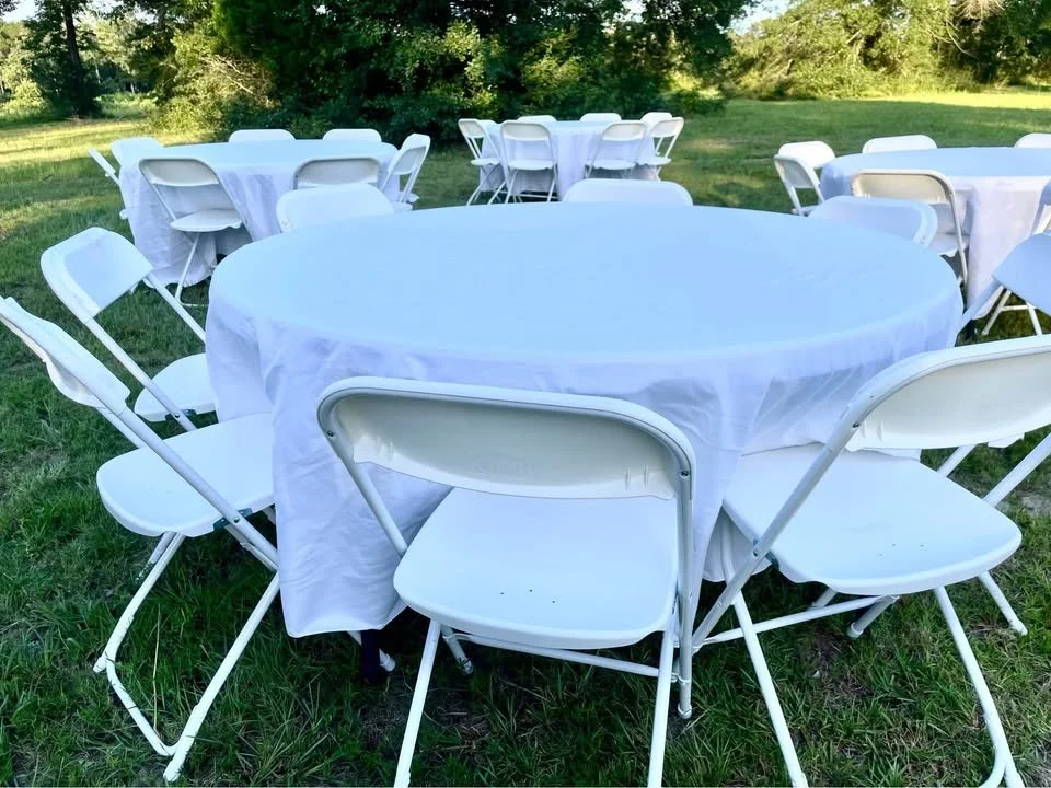 White round tables with white folding chairs set up on a grassy outdoor area, likely for an event or gathering.