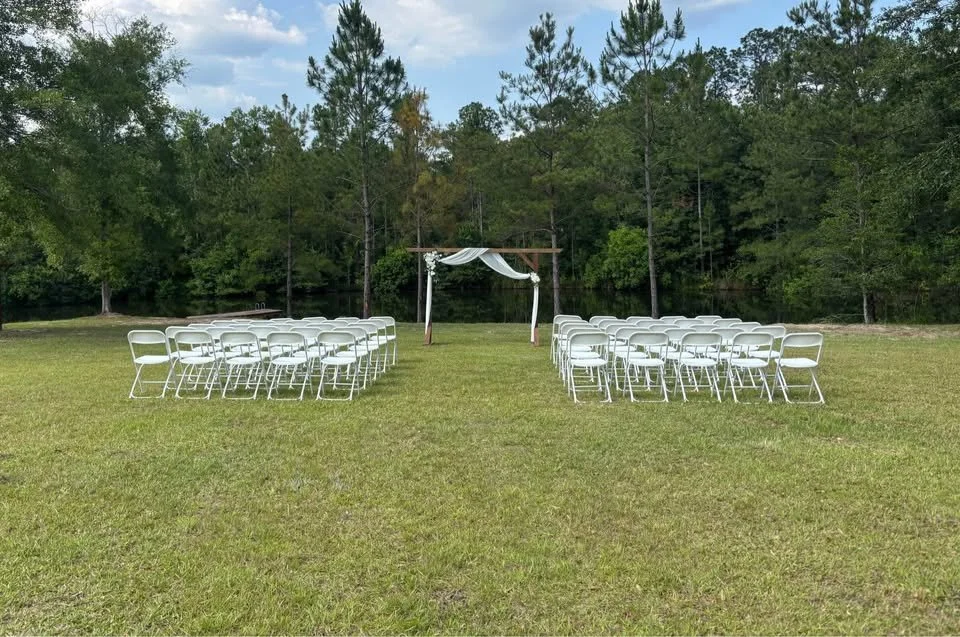 Chairs arranged in rows for an outdoor wedding ceremony with a wooden arch and white fabric drapery, set on a grassy field surrounded by trees.