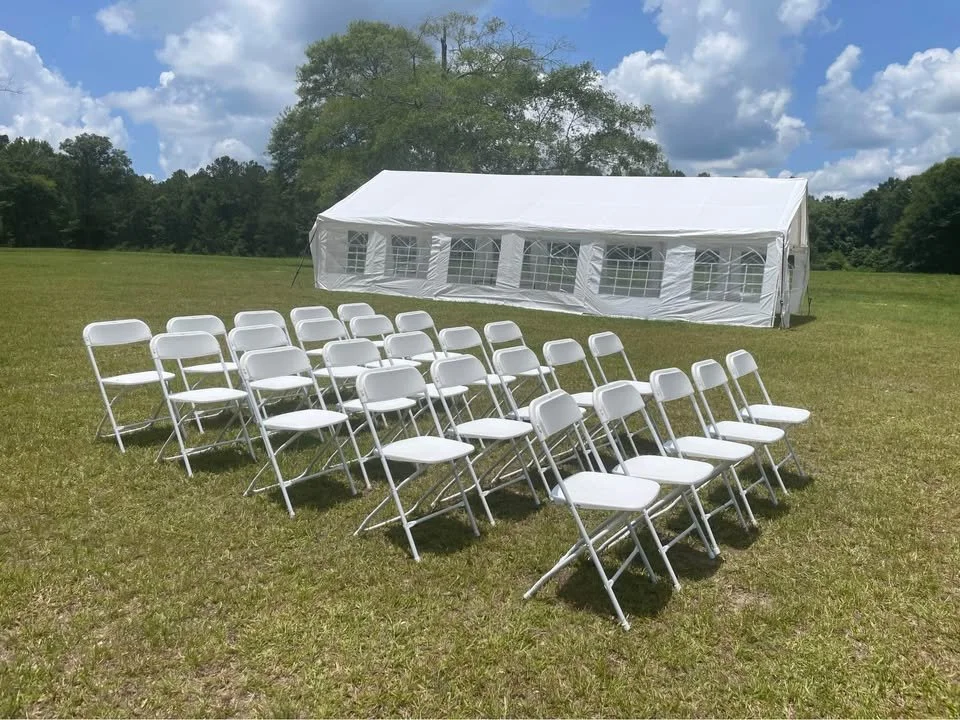 Empty white chairs arranged outdoors facing a large white event tent in a grassy field with trees and partly cloudy sky in the background.