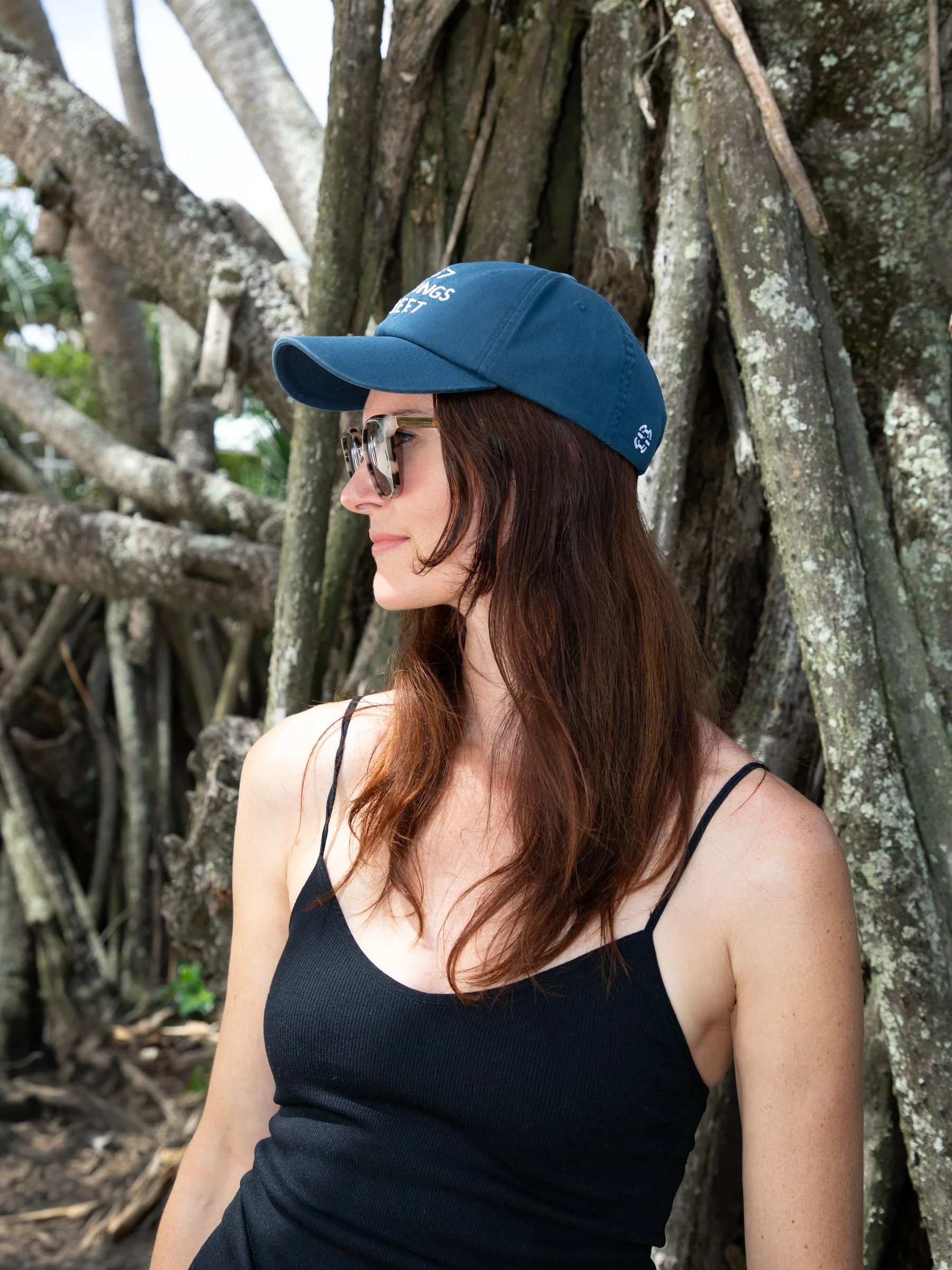 A woman with long brown hair wearing sunglasses, a navy blue cap, and a black spaghetti strap top, standing outdoors against a large tree with sprawling roots.
