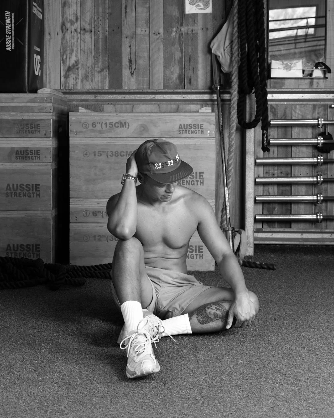 A shirtless young man sitting on the gym floor with one knee up, wearing shorts, sneakers, and a cap, in a gym setting with wooden boxes and workout equipment behind him.