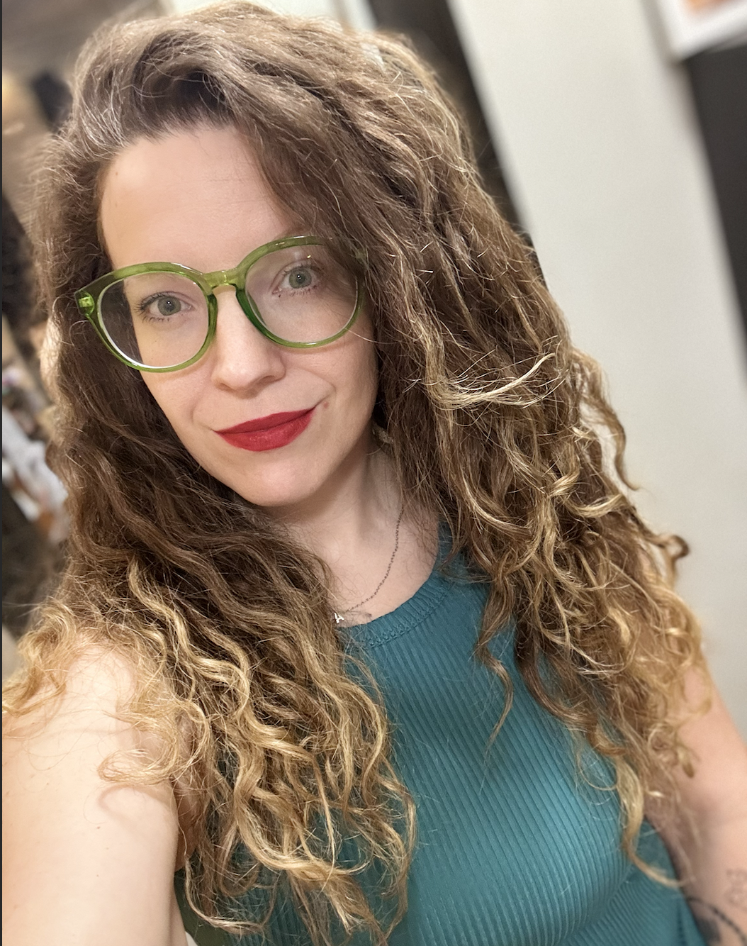 A woman with curly brown hair and green glasses, wearing red lipstick and a teal top, taking a selfie indoors.