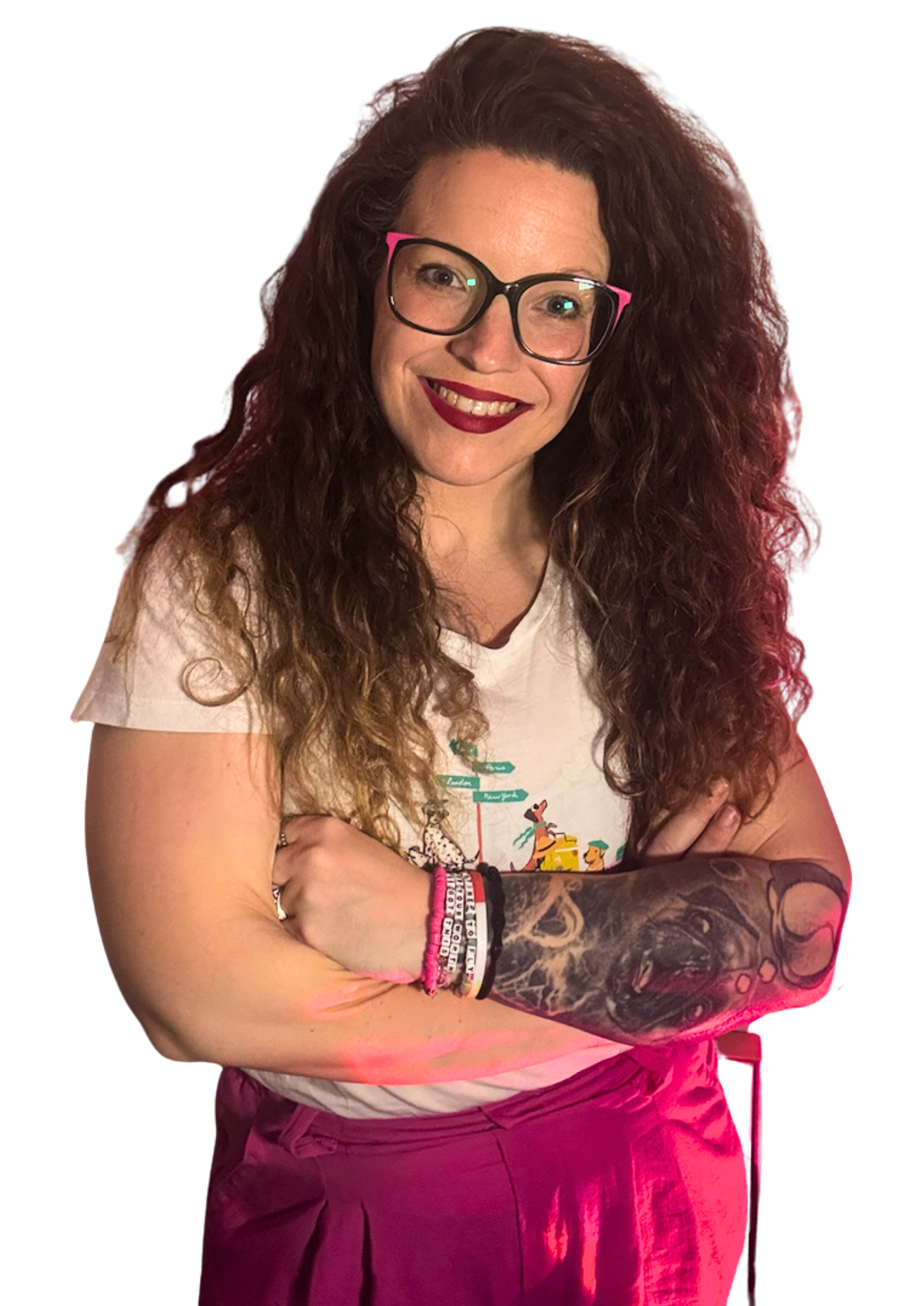 A woman with long, curly hair, glasses, and tattoos on her right arm, smiling and crossing her arms, wearing a white T-shirt and maroon pants.