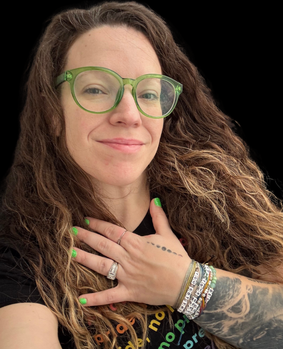 A woman with long curly hair wearing green glasses, showing a gentle smile, with her hand over her chest, displaying colorful bracelets, tattoos, and manicured nails matching her glasses.