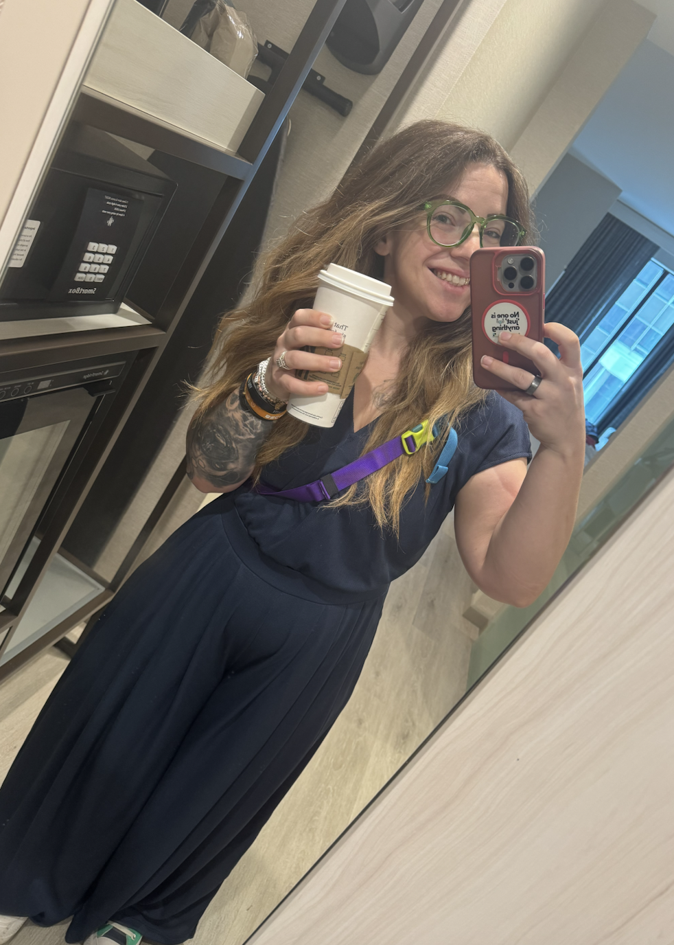 Woman in navy scrubs holding a coffee cup taking a selfie in a hotel room mirror.