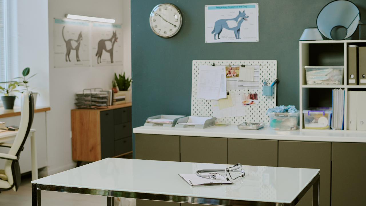 Medical office with a white clock on a green wall, a stethoscope and clipboard on a white table, and cabinets with office supplies.