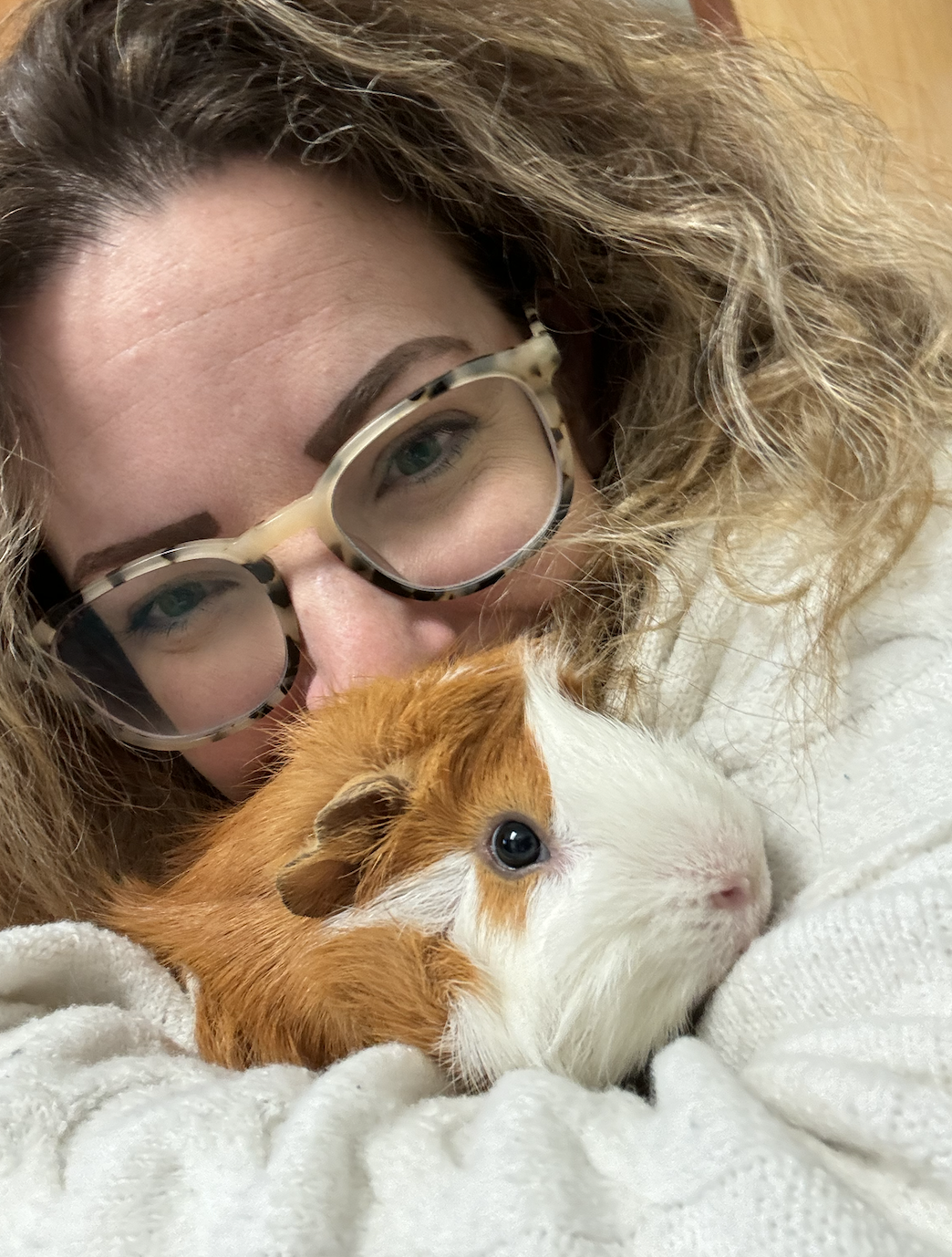 A woman with curly hair and glasses holding a guinea pig close to her face