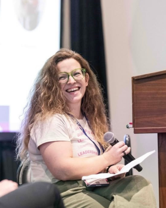 A woman with curly hair and green glasses smiling while holding a microphone and a piece of paper, sitting at a table during a presentation or event.