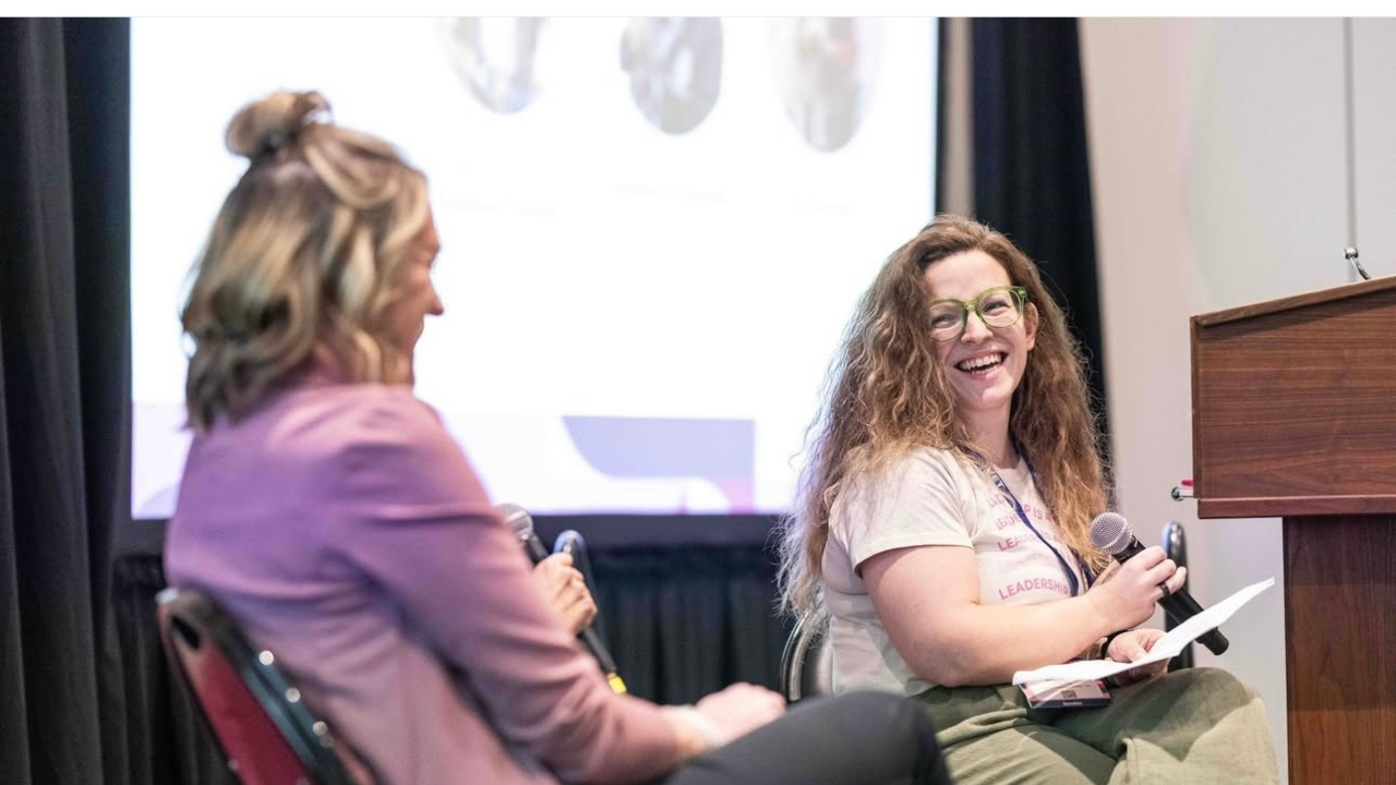 Two women sitting and conversing at a panel discussion or conference, with one woman smiling and holding a microphone, the other woman blurred in the foreground.