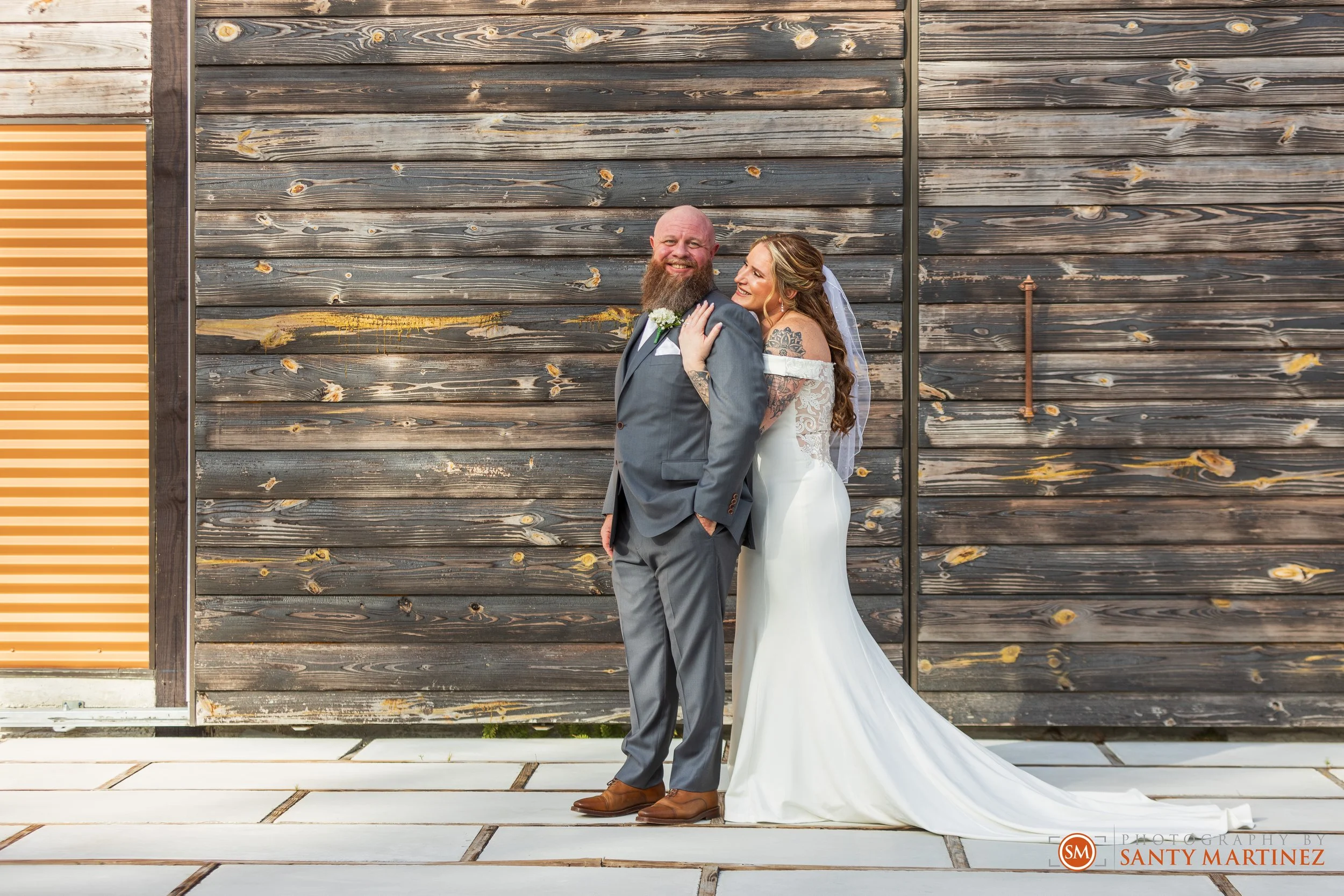 Bride in white dress and veil poses with groom in gray suit in front of wooden wall.