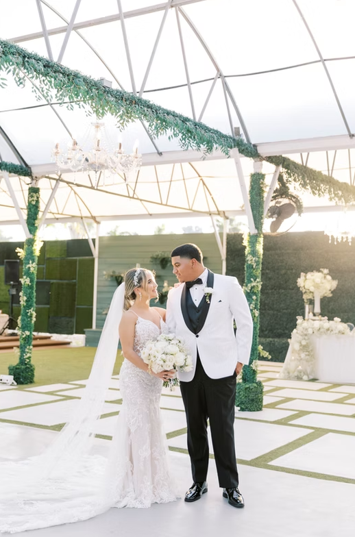 Bride and groom smiling at each other under an elegant wedding canopy with floral decorations and a chandelier.