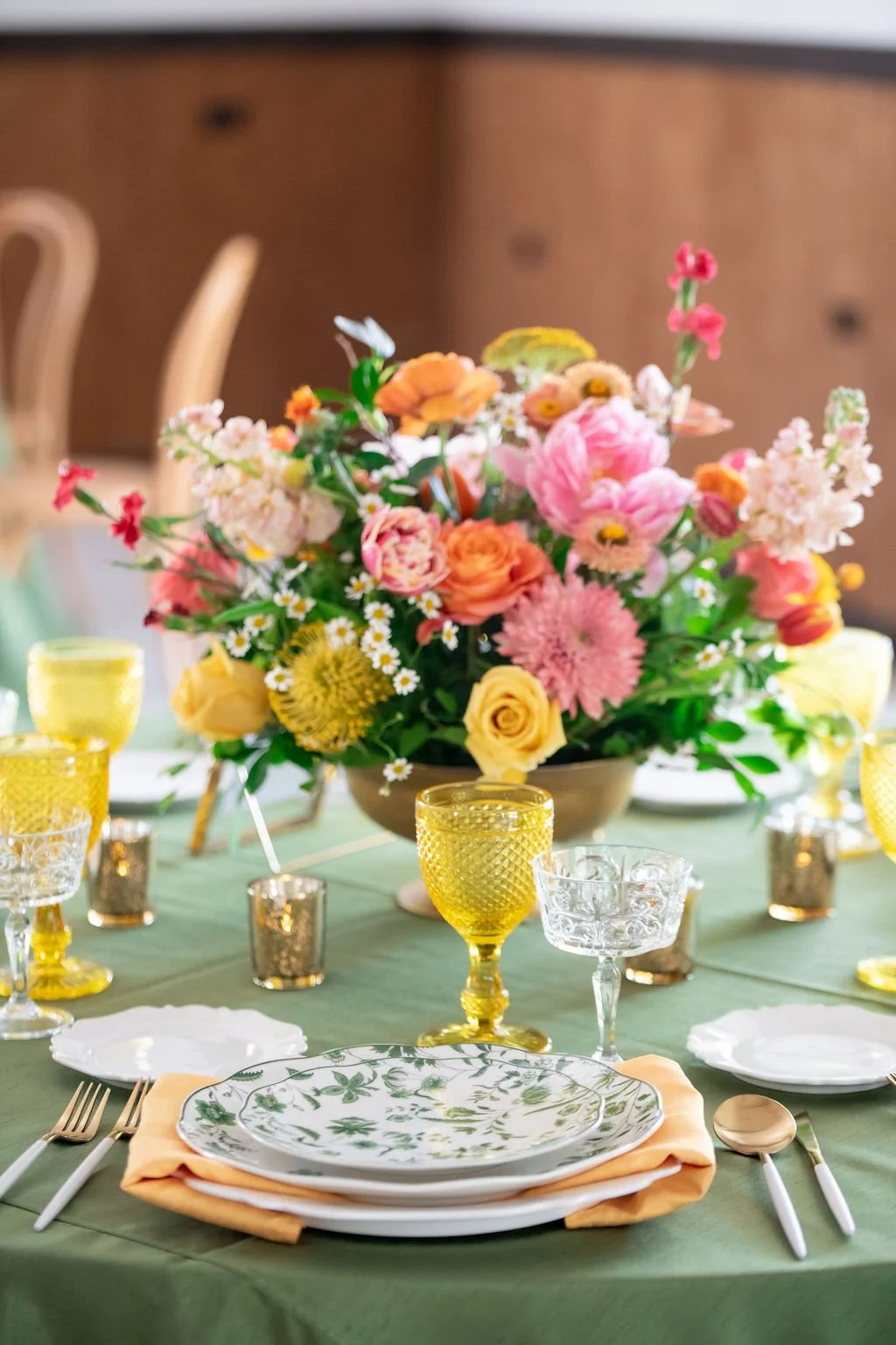 Elegant table setting with floral centerpiece, green tablecloth, patterned plates, yellow and clear glassware, and gold utensils.