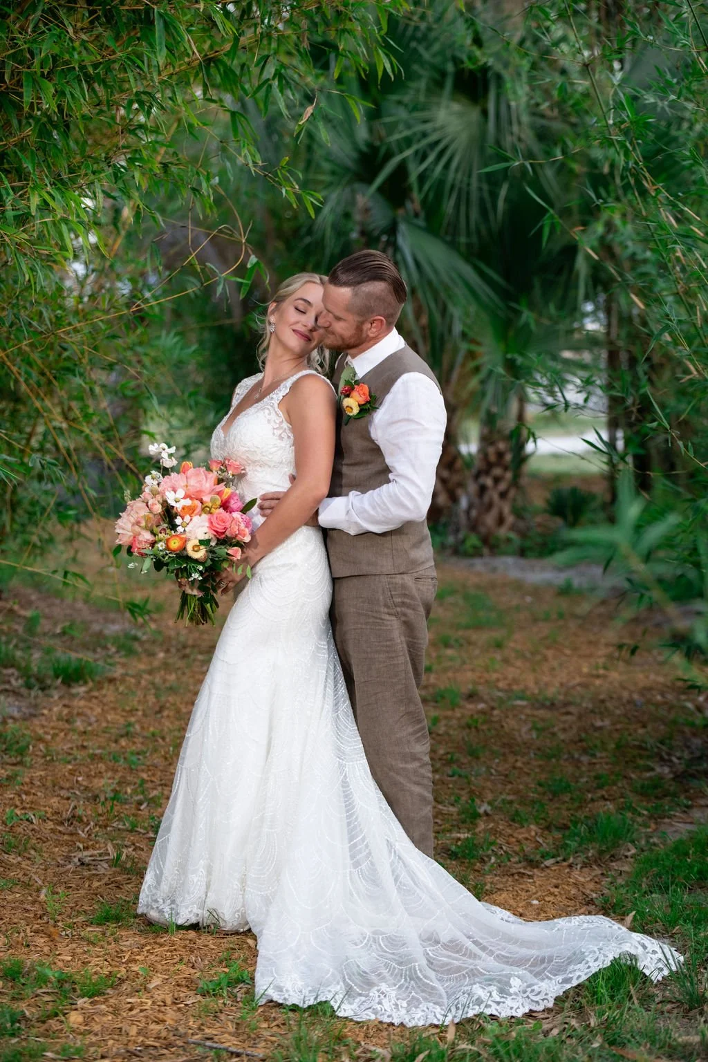 A bride and groom embrace in an outdoor setting surrounded by greenery. The bride is wearing a white lace wedding dress and holding a bouquet of colorful flowers. The groom is dressed in a brown suit with a white shirt, and has a boutonniere matching