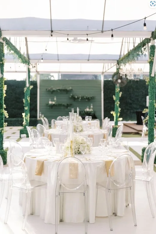 Elegant outdoor wedding reception setup with round tables, white tablecloths, clear chairs, and floral centerpieces under a transparent tent.