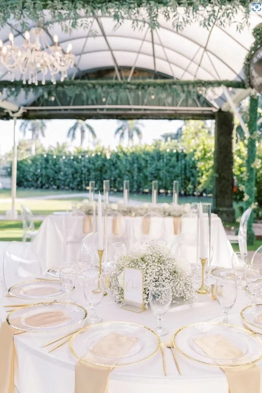 Elegant outdoor wedding reception setup with a decorated table featuring white linens, gold accents, glassware, and floral centerpieces under a canopy with greenery and a chandelier.