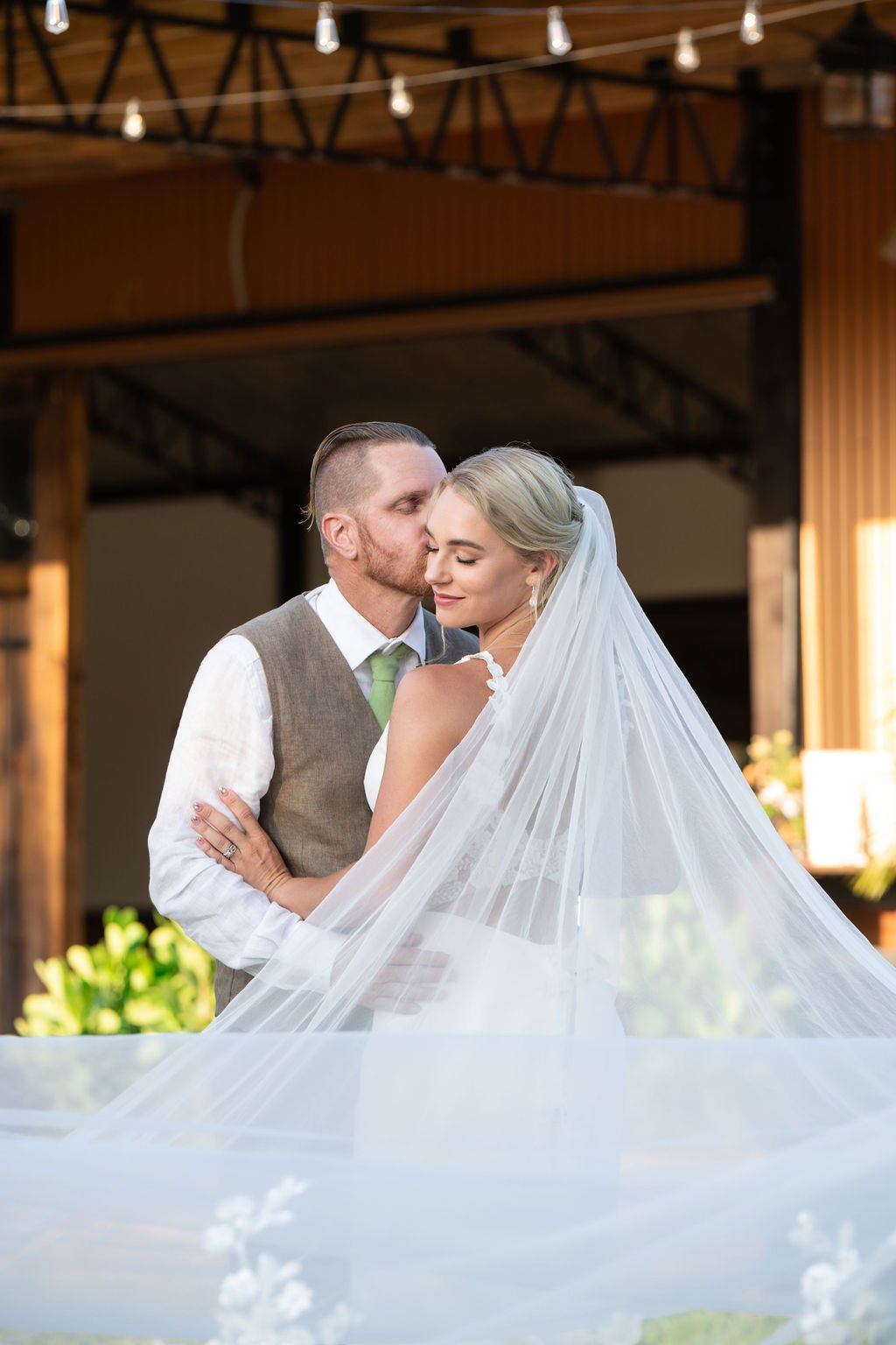Bride and groom embracing, with the groom kissing the bride's forehead, both wearing wedding attire, in an outdoor setting.