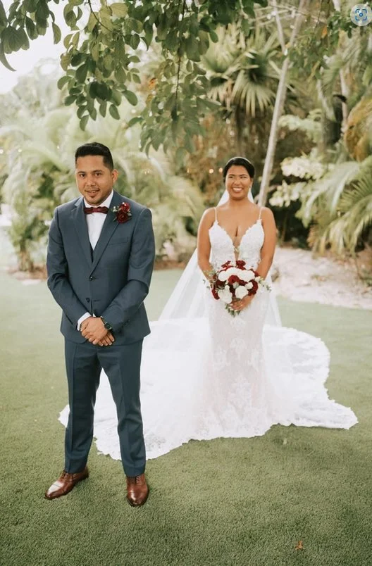 Bride and groom posing outdoors on grass with greenery background. The bride is in a lace wedding dress holding a bouquet, and the groom is in a blue suit with a red bow tie.