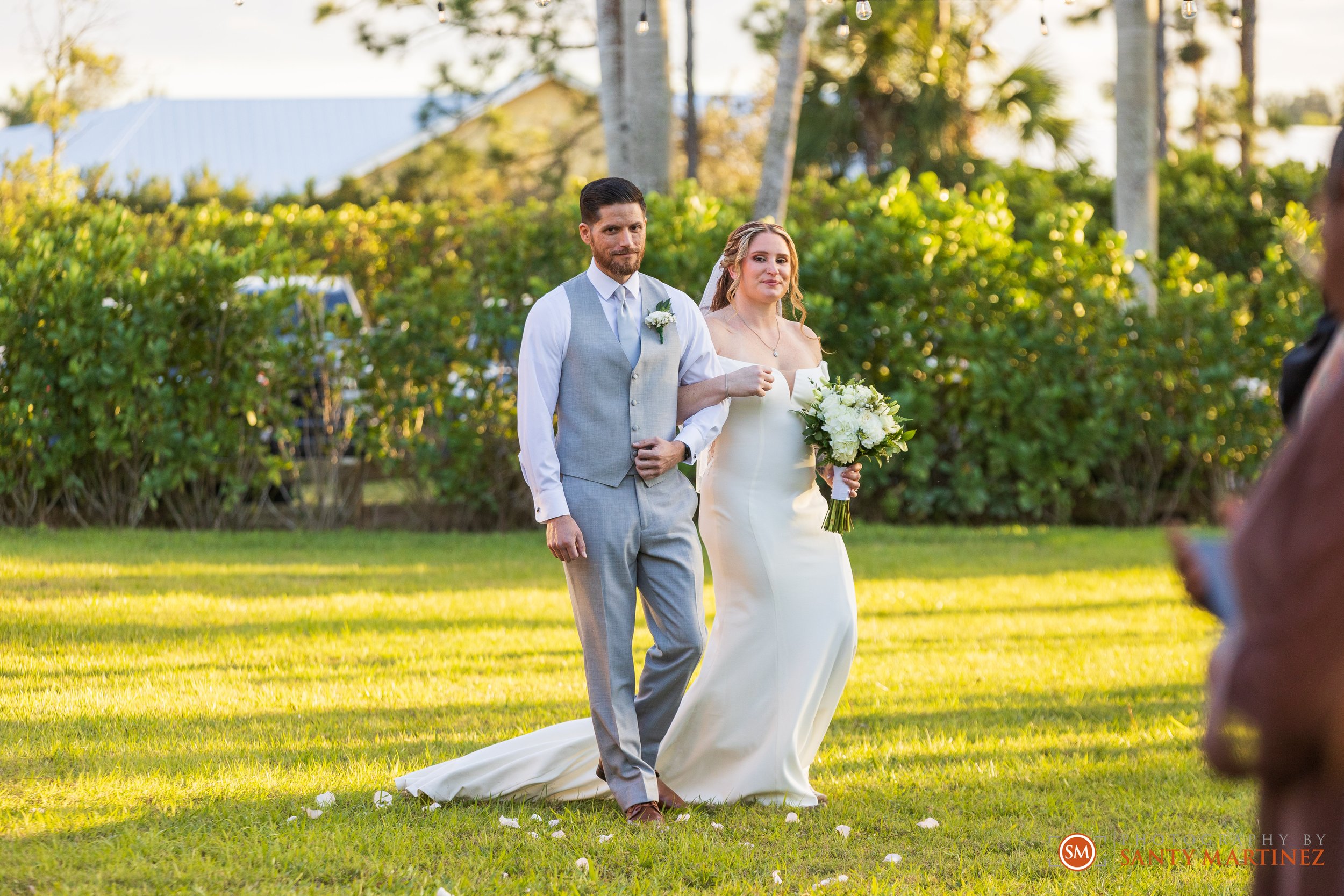 Bride and groom walking on grass in outdoor wedding ceremony, bride holding bouquet, sunny background with trees.