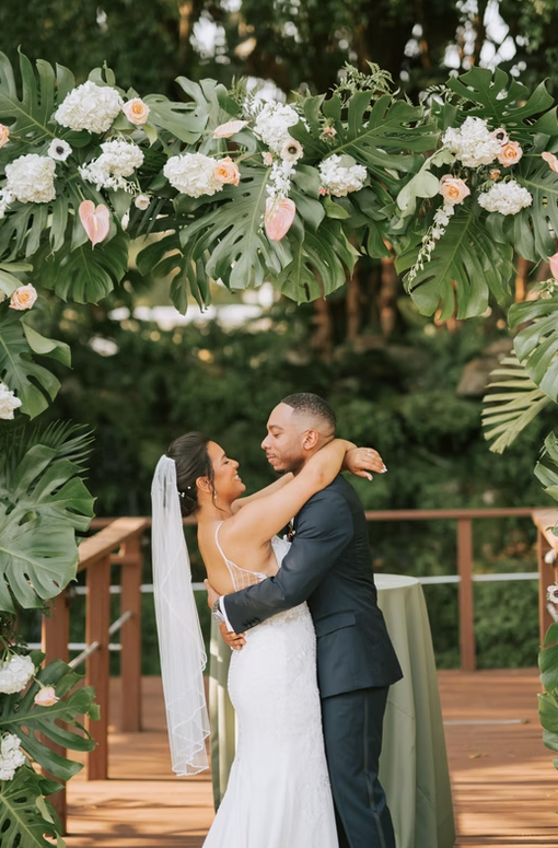 Bride and groom embracing under floral arch at outdoor wedding ceremony.