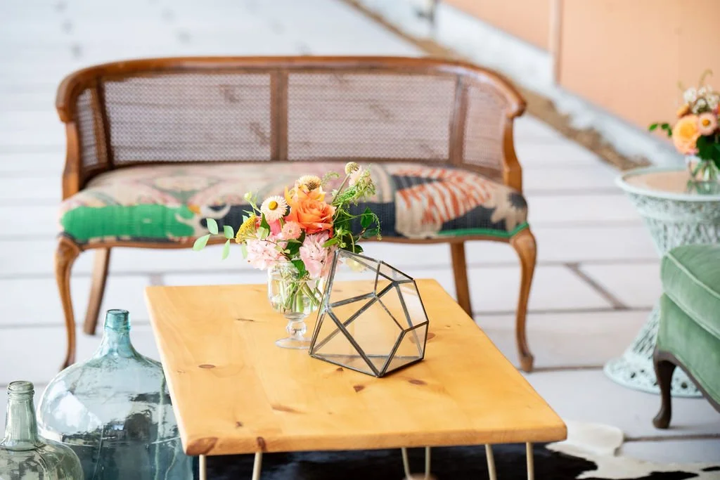 Vintage loveseat with patterned cushion, wooden coffee table with geometric decor, floral arrangement, and glass bottle in an eclectic interior setting.