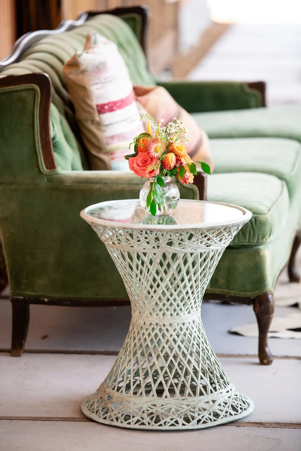 Elegant green velvet sofa with decorative pillows beside a white wicker side table holding a glass vase filled with colorful flowers.