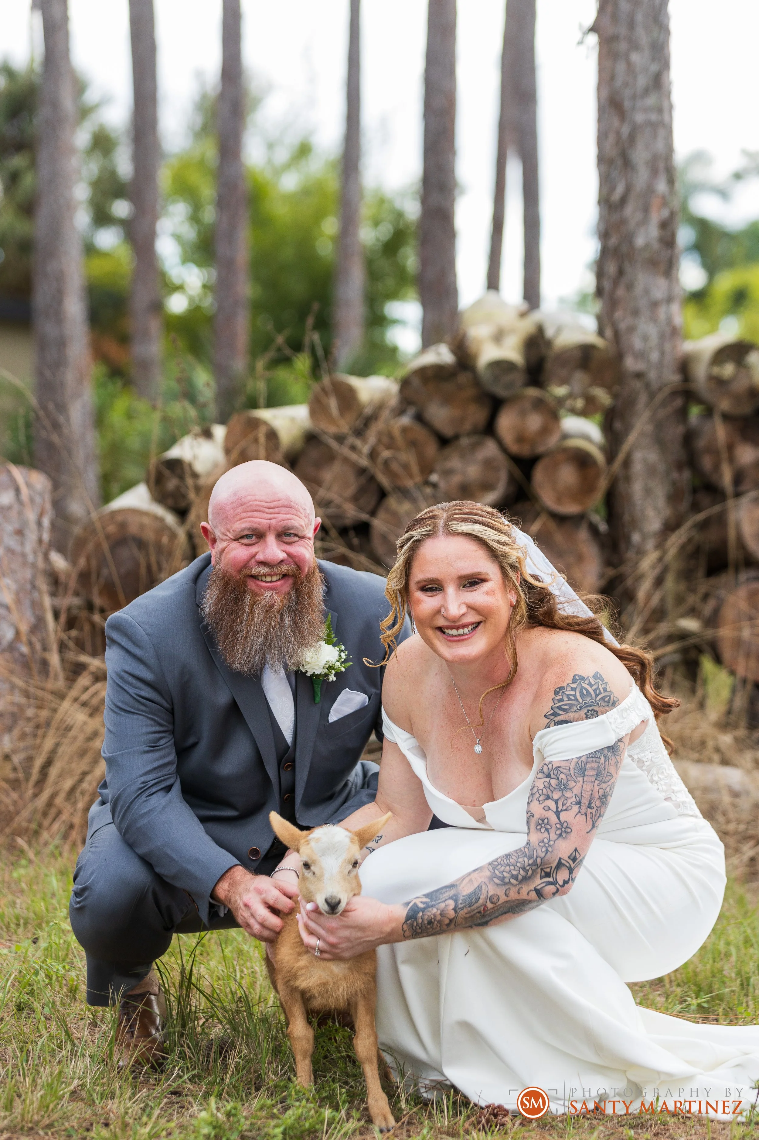 Smiling bride and groom outdoors with a small goat in a wooded area.