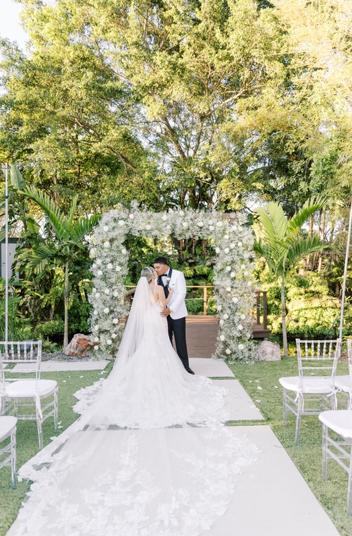 Bride and groom kissing under a floral arch at an outdoor wedding ceremony with chairs and green foliage.