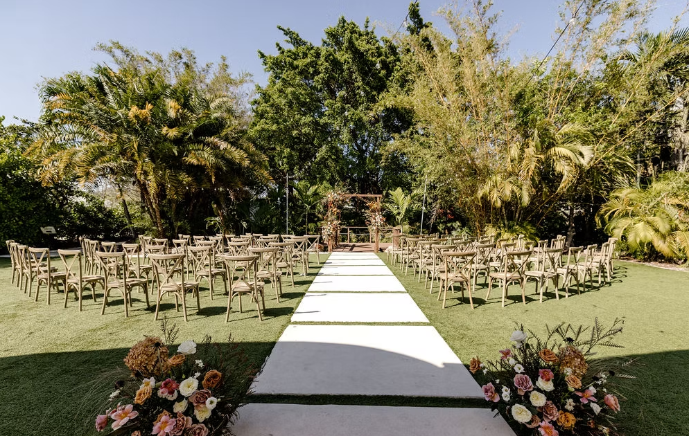 Outdoor wedding ceremony setup with wooden chairs and floral decorations on a grassy area, surrounded by trees and plants.