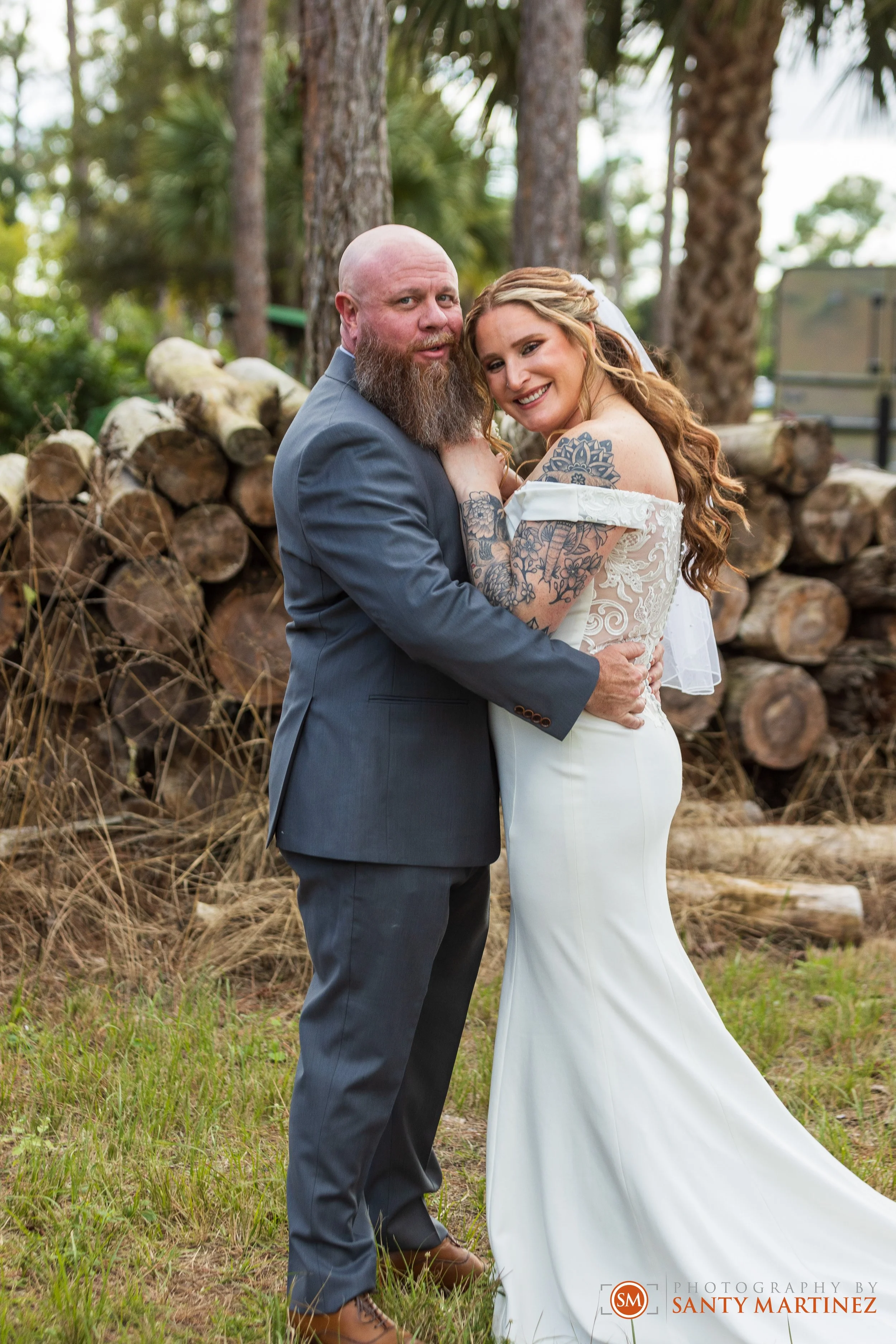 A couple posing for their wedding photo outdoors with trees and logs in the background. The groom is in a suit, and the bride is in a white dress, showing her tattooed arm.