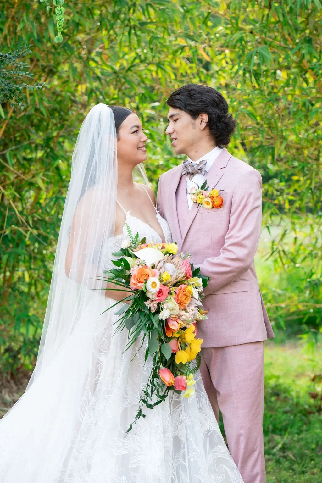 A bride and groom standing together outdoors, with the bride wearing a white wedding dress and veil, and holding a colorful bouquet of flowers. The groom is wearing a light pink suit with a floral boutonniere. They are surrounded by greenery.