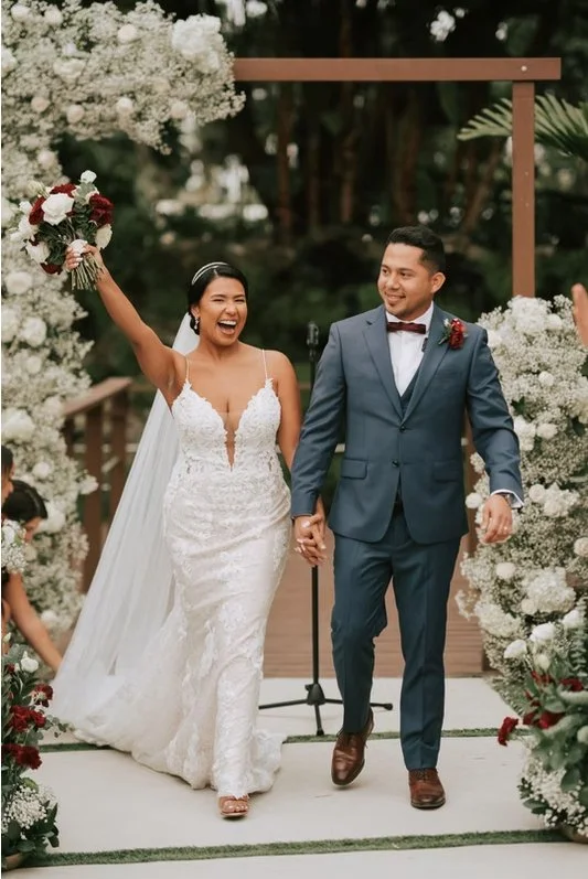 Bride and groom walking hand in hand under a floral arch at their wedding, bride holding bouquet.
