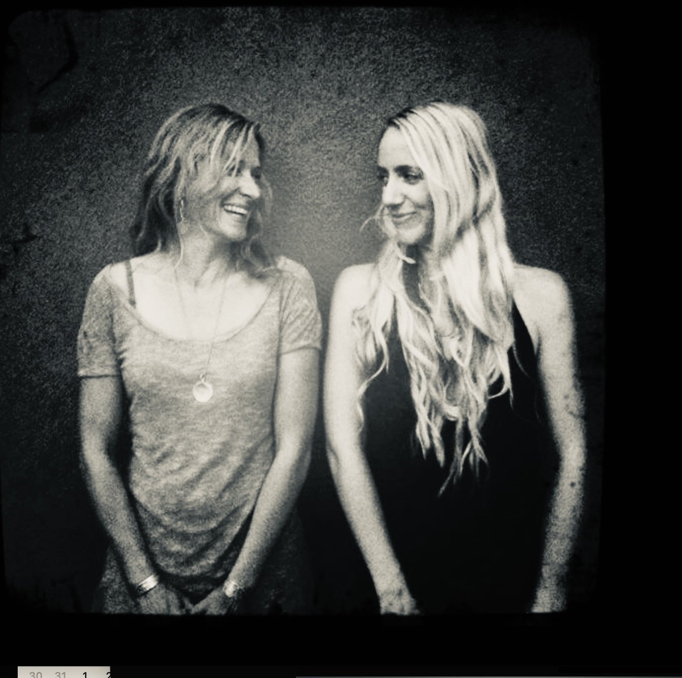 Two women standing close together, one with short wavy hair smiling and the other with long wavy hair looking at her, both in casual outfits, in black and white.