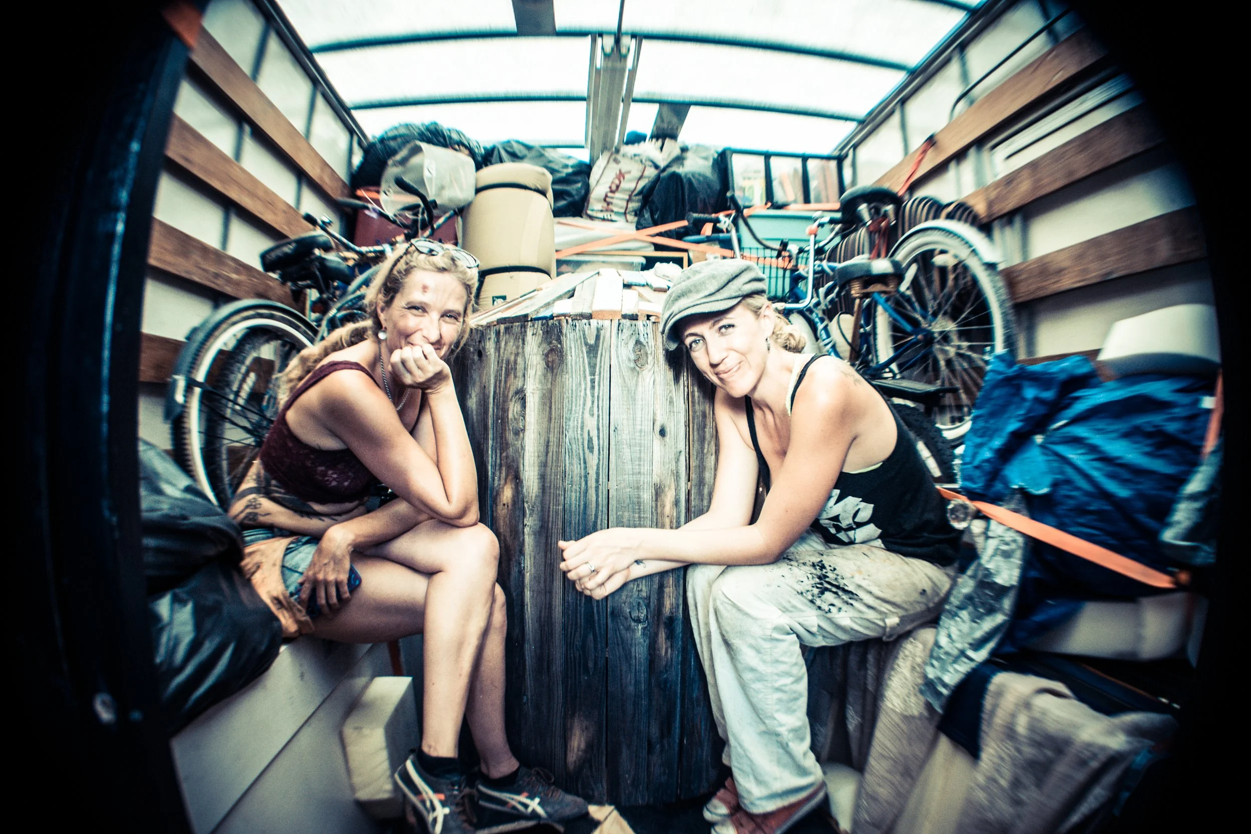 Two women sitting inside a box truck with packed belongings and bicycles around them, smiling at the camera.