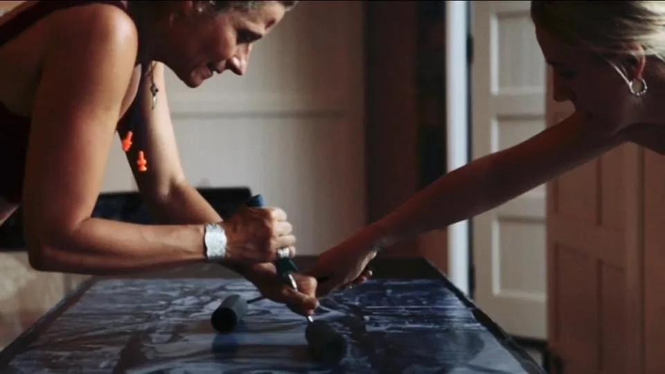 Two women are leaning over a table, sealing one of their murals. They are both applying sealant with tools and brushes, stretching out over the work. The scene appears to be in a home setting with warm lighting.