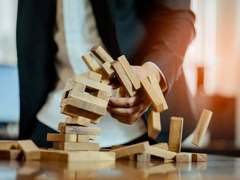 A person in business attire playing Jenga, a wooden block stacking game, with some blocks falling.