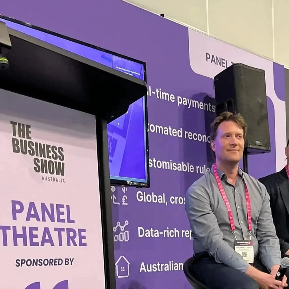 Roger sitting on stage at a panel discussion during The Business Show Australia with a crowd in the background and purple banners with white text.