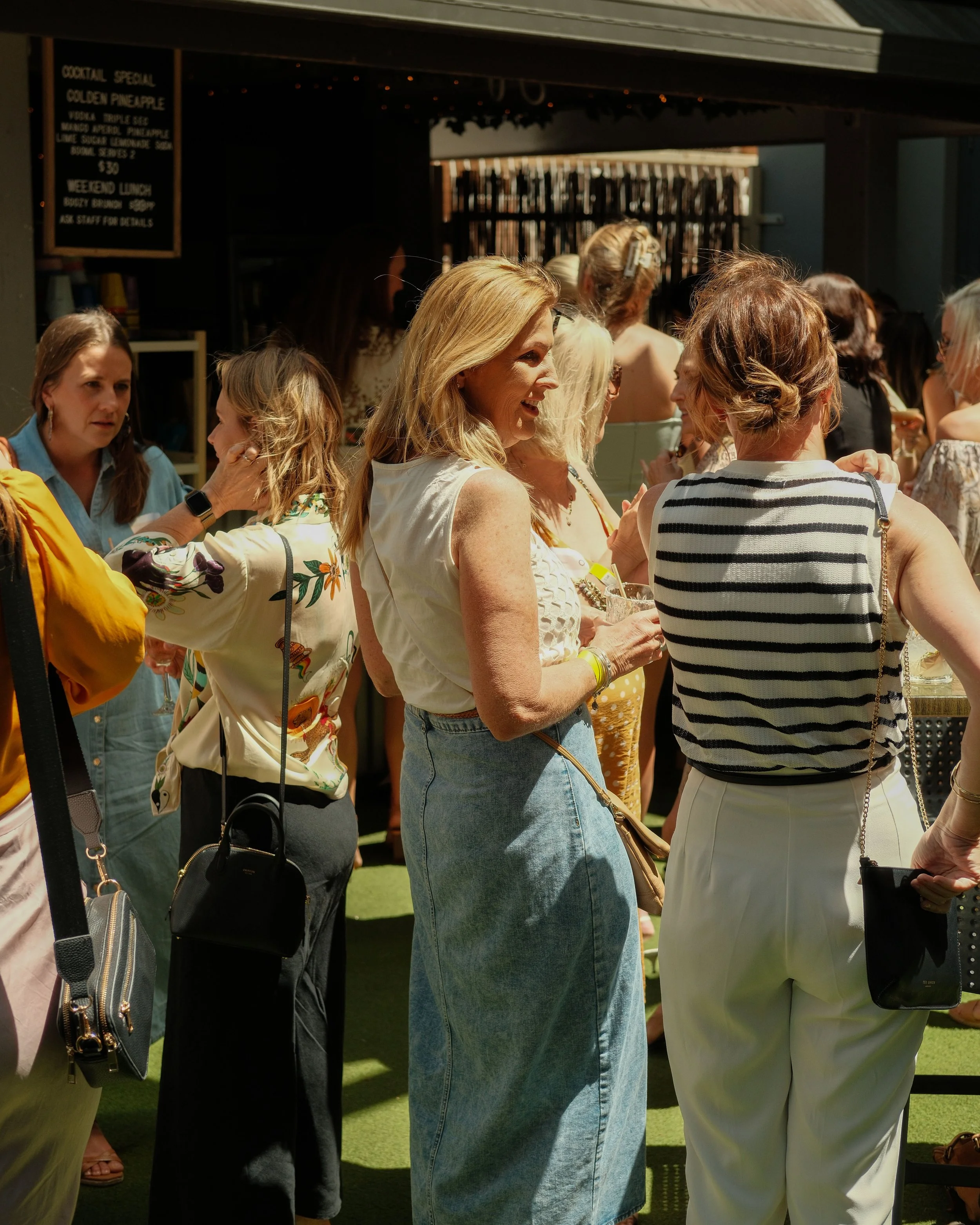A group of women socializing outdoors at a daytime event, some holding drinks, with a bar or food stand in the background.