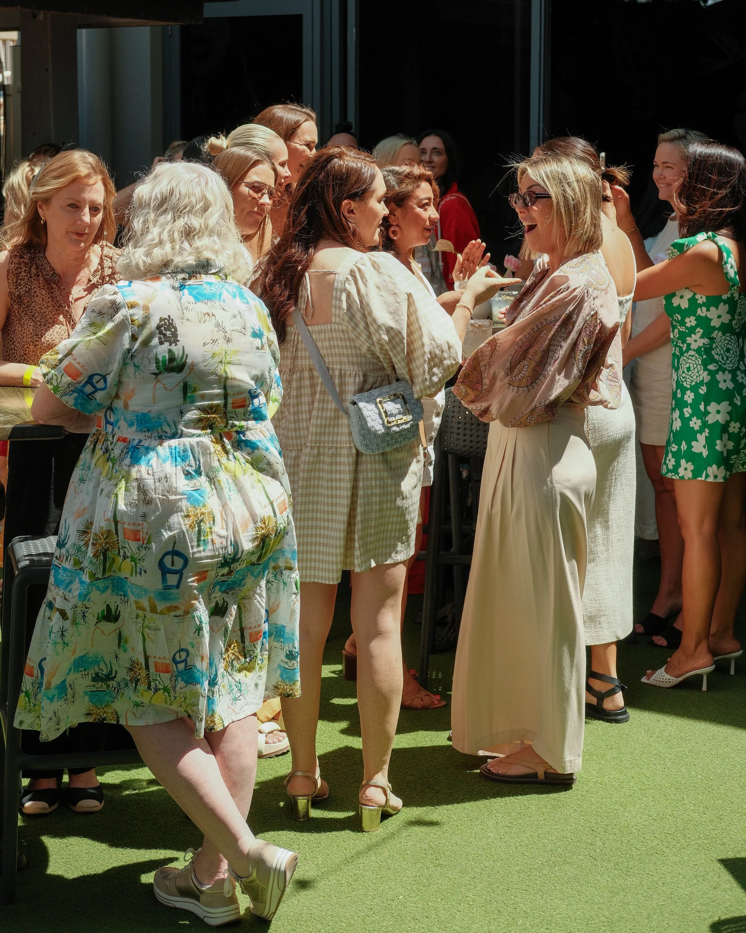 Group of women socializing at an outdoor gathering, standing on green artificial grass, with some wearing light-colored or floral outfits, and smiling.