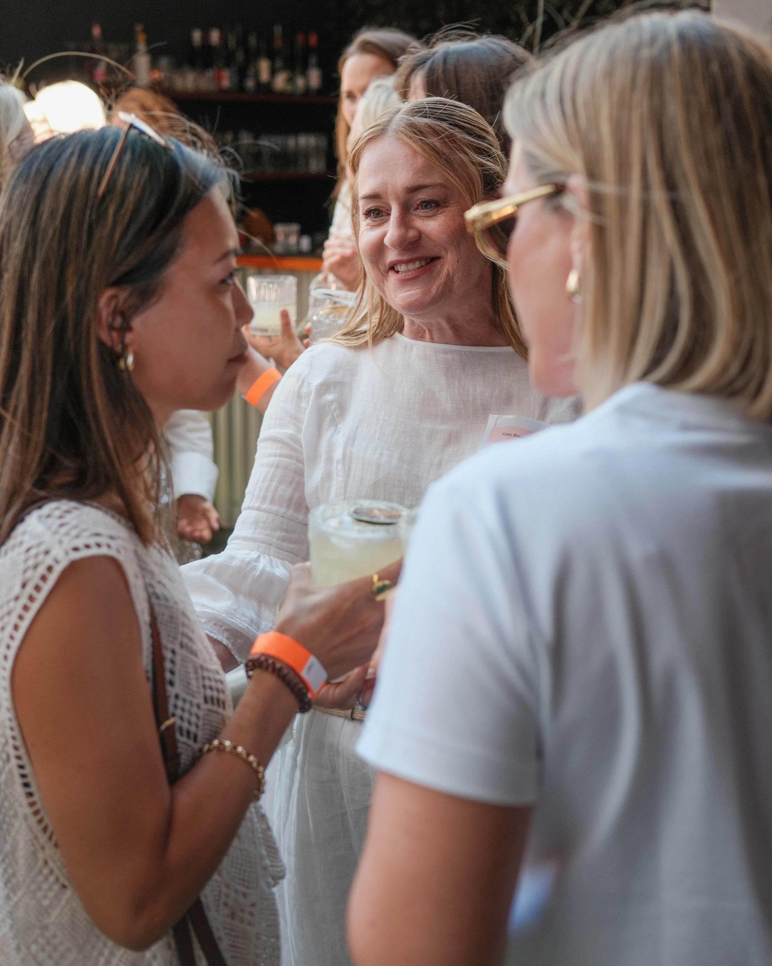 A group of women at a social gathering, engaging in conversation and holding drinks, with a bar in the background.